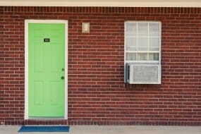 Image shows an ac window unit in a brick building with a green door