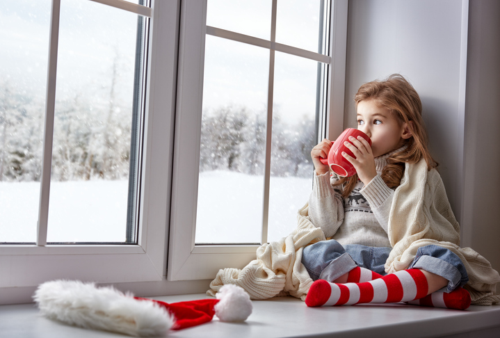 little girl sitting by the window with a cup of hot drink and looking at the winter forest