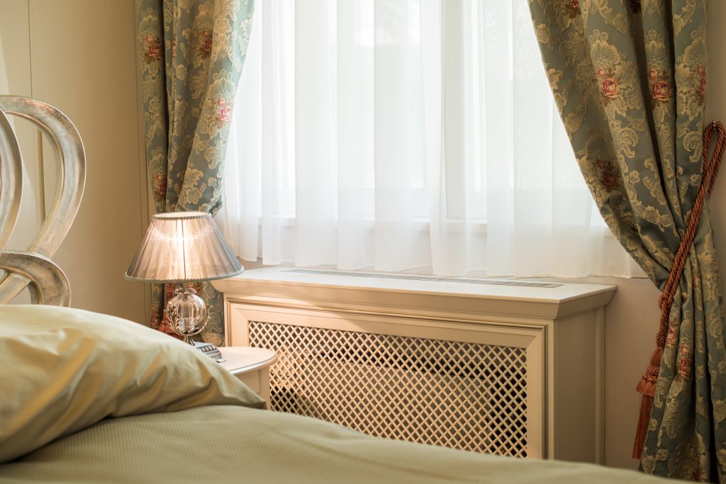 Image of a radiator in a bedroom underneath a window with a decorative, cream-colored cover.