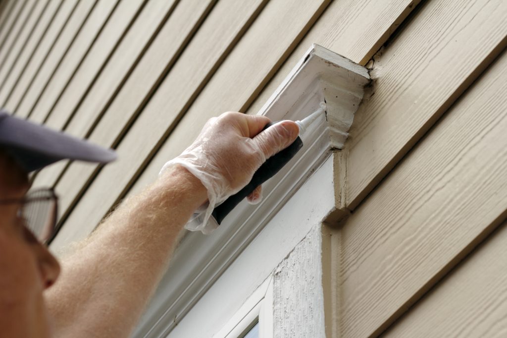 A close-up image of a person applying window sealant to the exterior trim of a window