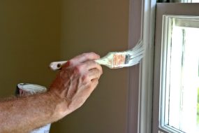 Close up of a man's arm and hand holding a paint brush and painting white latex paint on an interior window frame