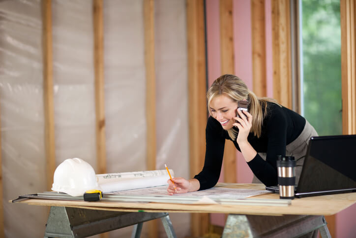 A young woman is looking at blueprints and talking on the phone. She is working at a construction site. Horizontally framed shot.