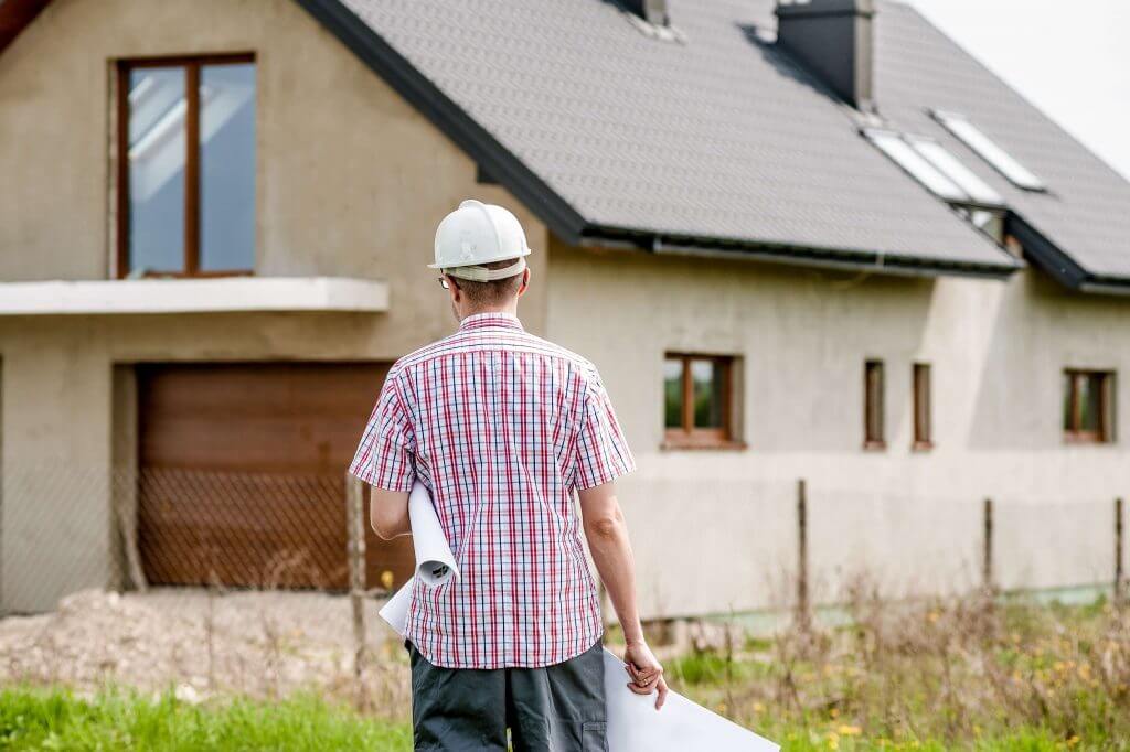 Image of a contractor from the back as they walk toward a home carrying plans and wearing a hard hat