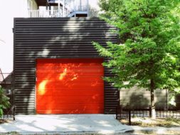 Orange, single car, roll-up garage door against dark building material with a tree and drive in front