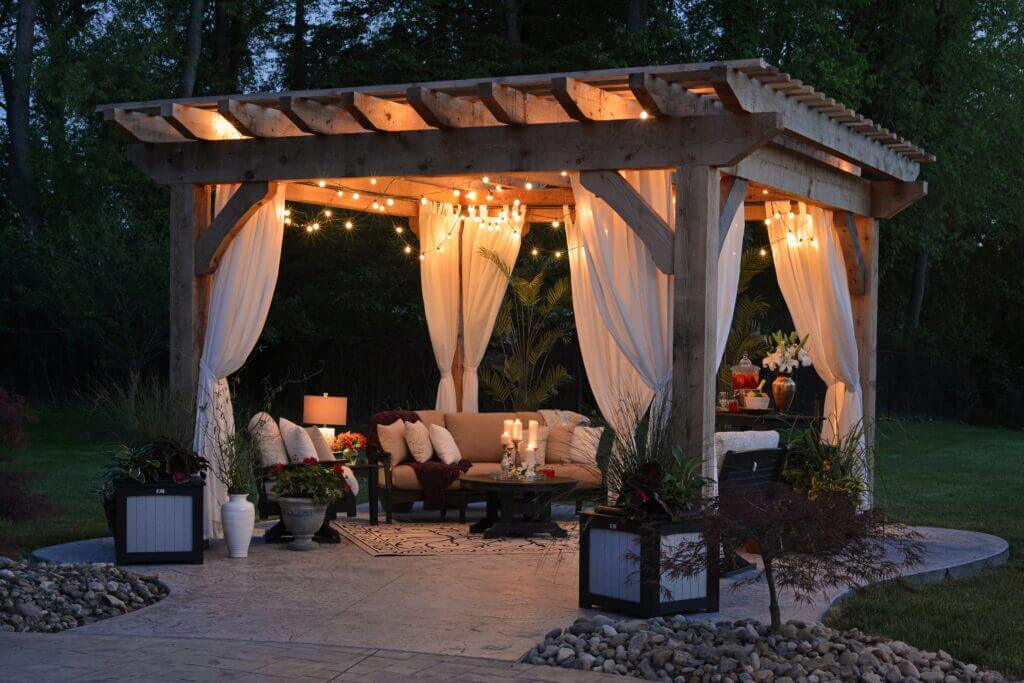 Image shows an outdoor seating area in the evening, with a pergola and lighting