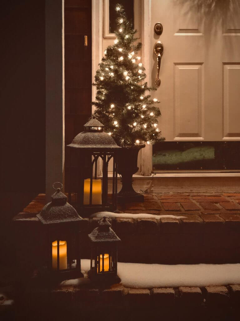 Christmas tree on a front porch with lanterns going up the stairs and snow across them
