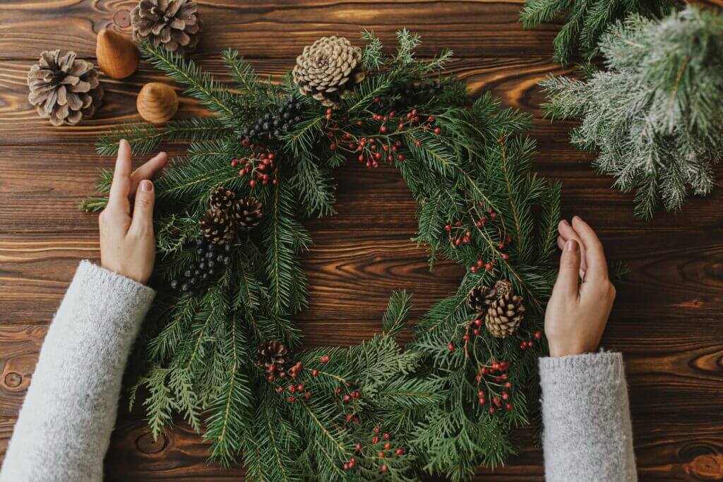 Closeup image of a wintery wreath with pinecones and berries and two hands putting it together