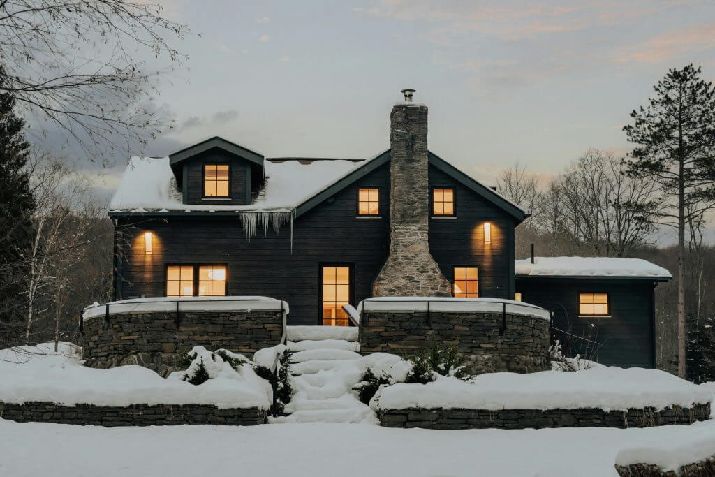 A photo of a black house with lights on in all the windows. Snow is covering the roof and ground.