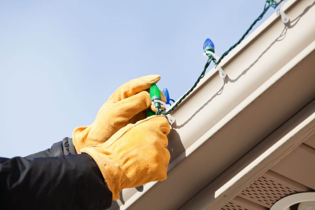 Gloved hands are installing a LED Christmas light onto a clip attached to a house rain gutter. 