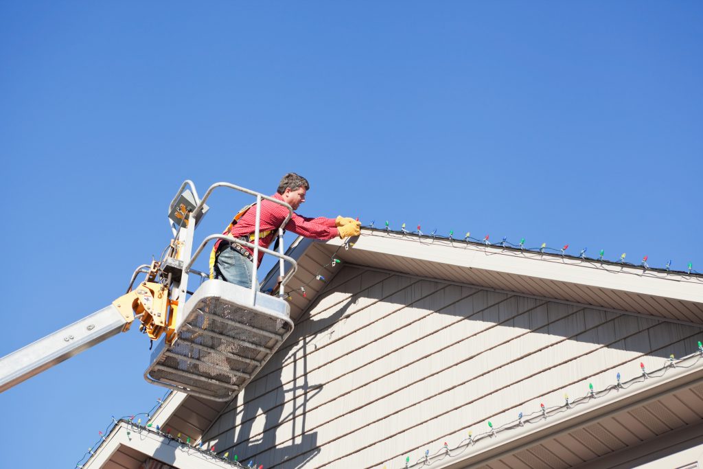 A professional man, standing in an aerial work platform with a safety harness, is installing LED Christmas lights to a house roof peak. 