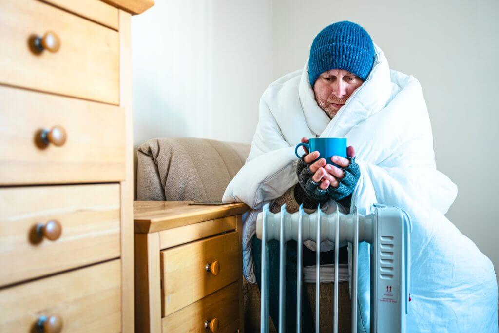 Color image depicting a mid adult man sitting next to an electric heater at home. He is cold and is wrapped up in a white duvet with a blue knit hat on his head.