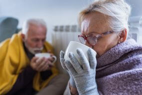 Senior couple covered with blanket sitting beside heater in living room, drinking hot tea from big cup and trying to warm in winter season and energy crisis
