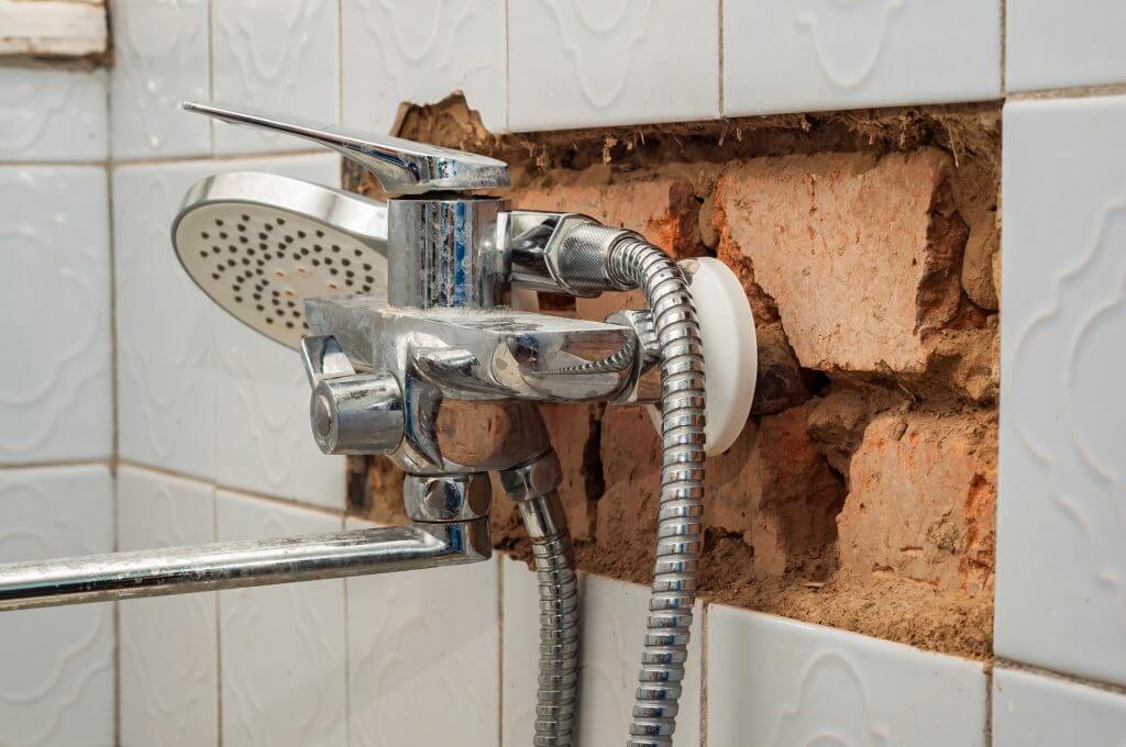 Close-up of a chrome shower faucet with a broken tiled wall showing the brick behind it, suggesting plumbing repairs or bathroom renovation is needed.