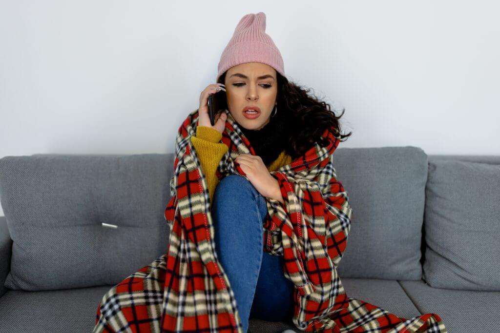 A woman in warm clothing is feeling cold and displeased while sitting at home and freezing and talking using a mobile phone.