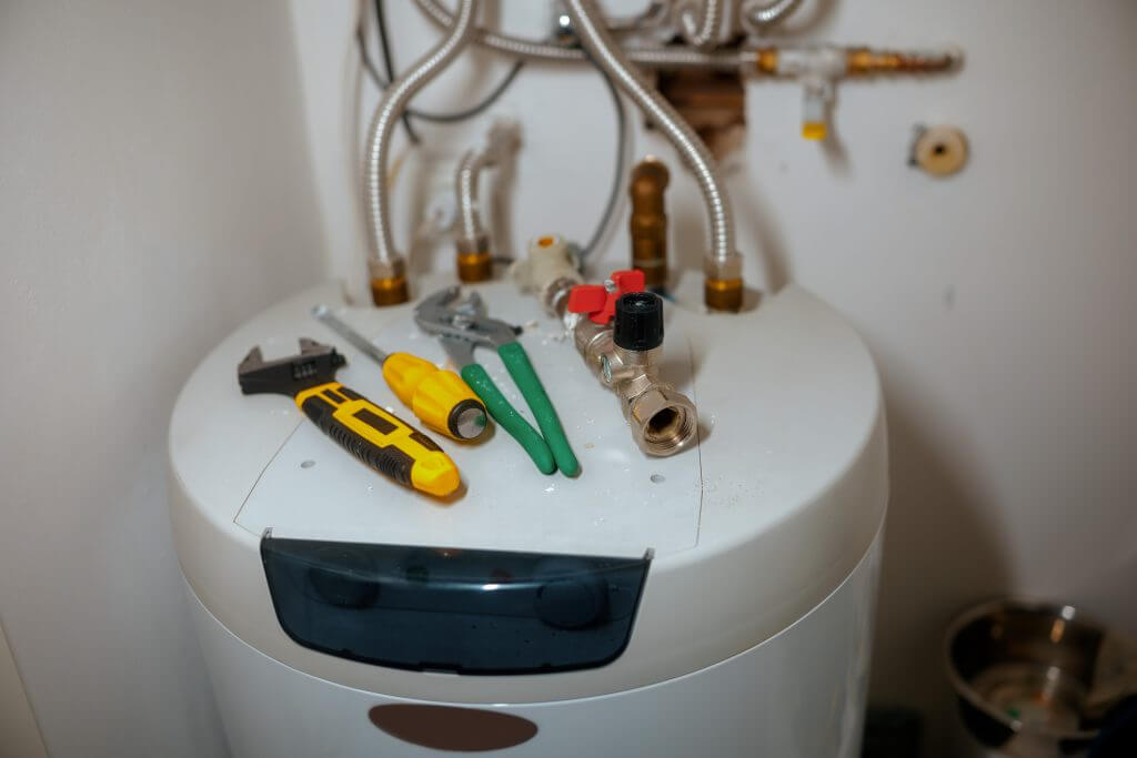 An adjustable wrench, screwdriver, and pliers rest on a white water heater, surrounded by pipes and valves, signaling active water heater repair