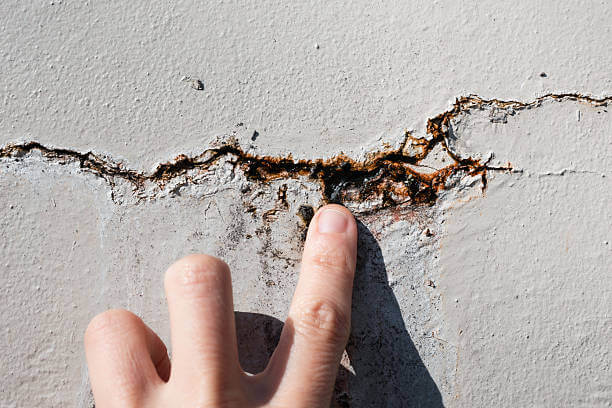 Close up of hand examining vertical crack in outdoor wall with rust and efflorescence. Repair foundation problems. 